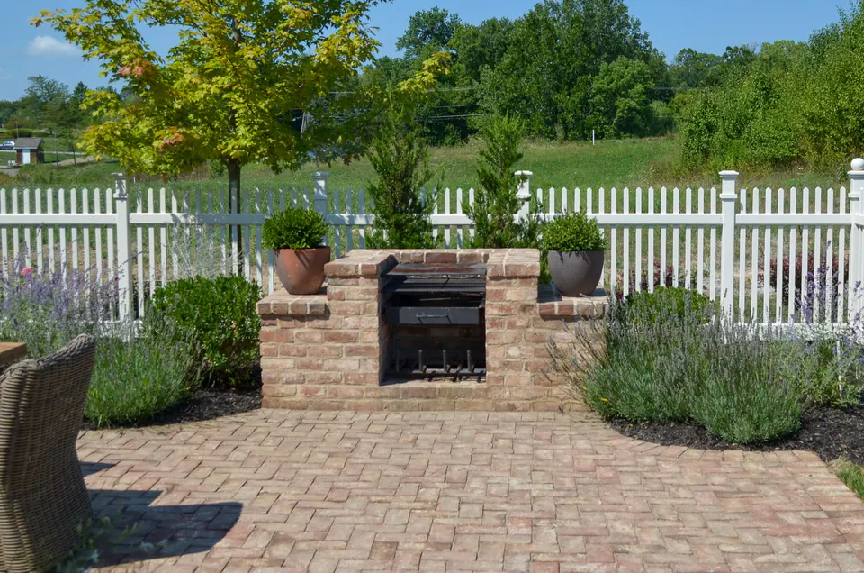 Brick outdoor grill station set into a paver patio with white picket fence, planters, and garden planting